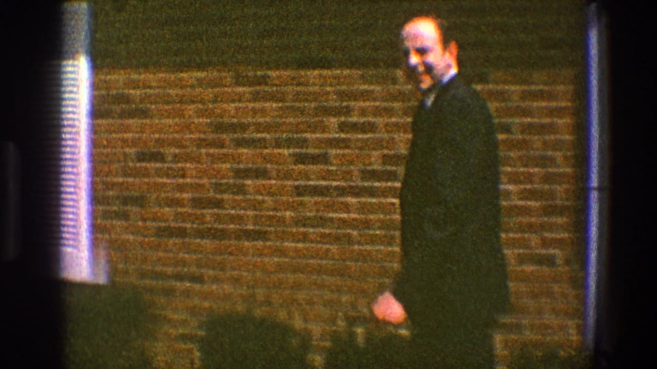 Man in a dark suit walking beside a brick house wall in daylight.