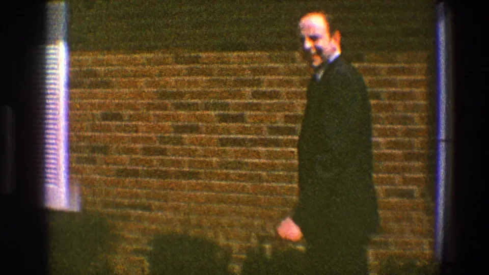 In 1969 Clarendon Hills home-movie footage, a suited man walks along a brick house exterior toward the camera.