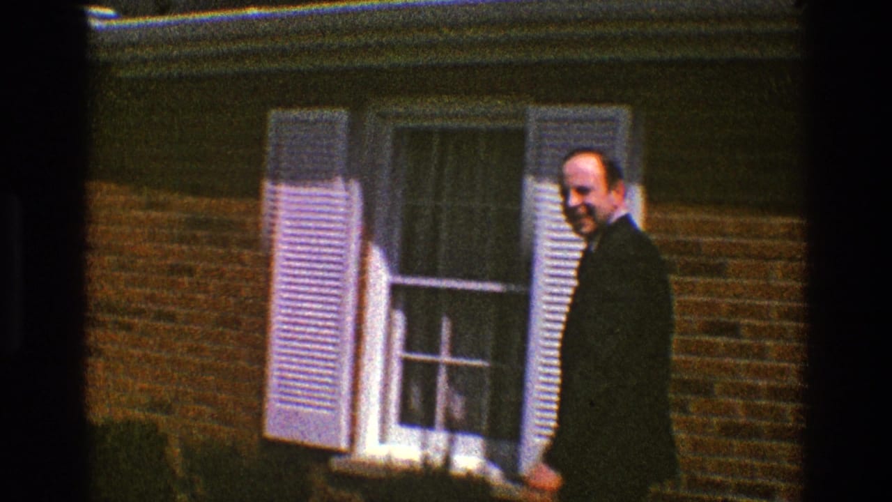 Man in a dark suit near white shutters and a window on a brick house.