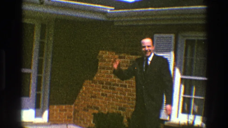 The man smiles and begins waving near a brick-and-shuttered suburban house exterior.