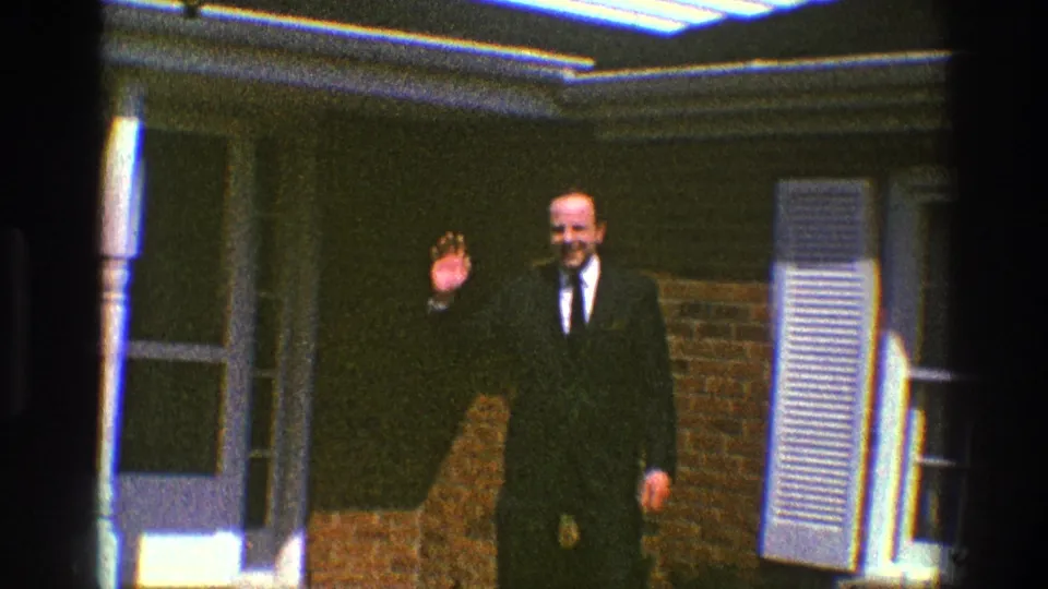 A suited adult man waves at the camera outside a brick home in 1969 Clarendon Hills footage.