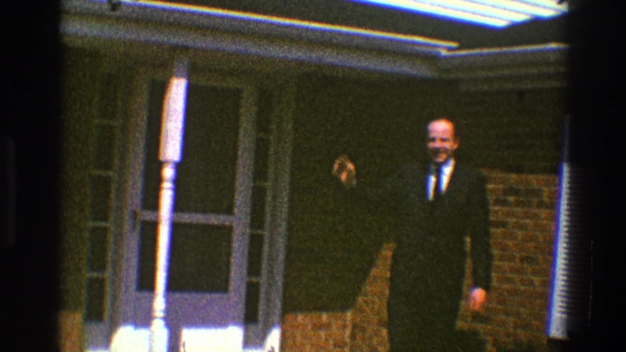 Suited man waves from a brick porch beside a white column and doorway.