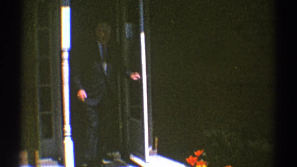 1969 Clarendon Hills home-movie frame with a man in a dark suit and tie opening or standing beside a white screen door on a porch.