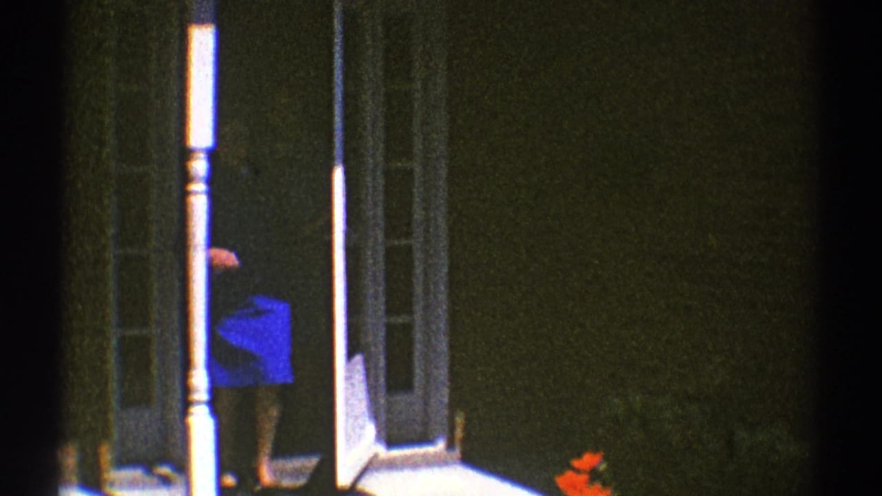 Woman in a blue outfit stands partly inside a house doorway beside white porch posts.