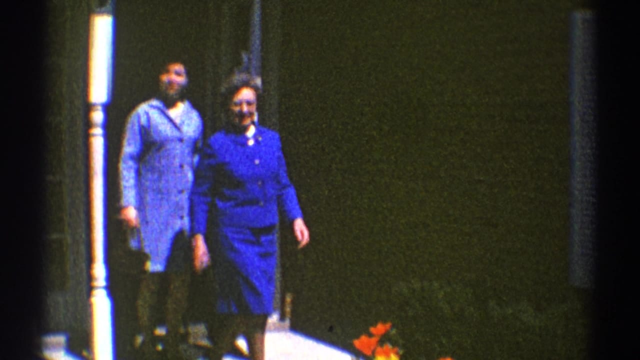 Two women in blue outfits walk out from a house entrance toward the porch.
