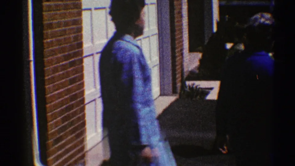 The women move along the garage-front walkway, with the darker blue outfit partly obscured by shadow and the light coat near the door.