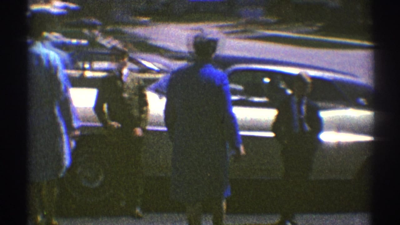 Several adults gather near the side of a parked sedan, partly obscured by film gate edges.