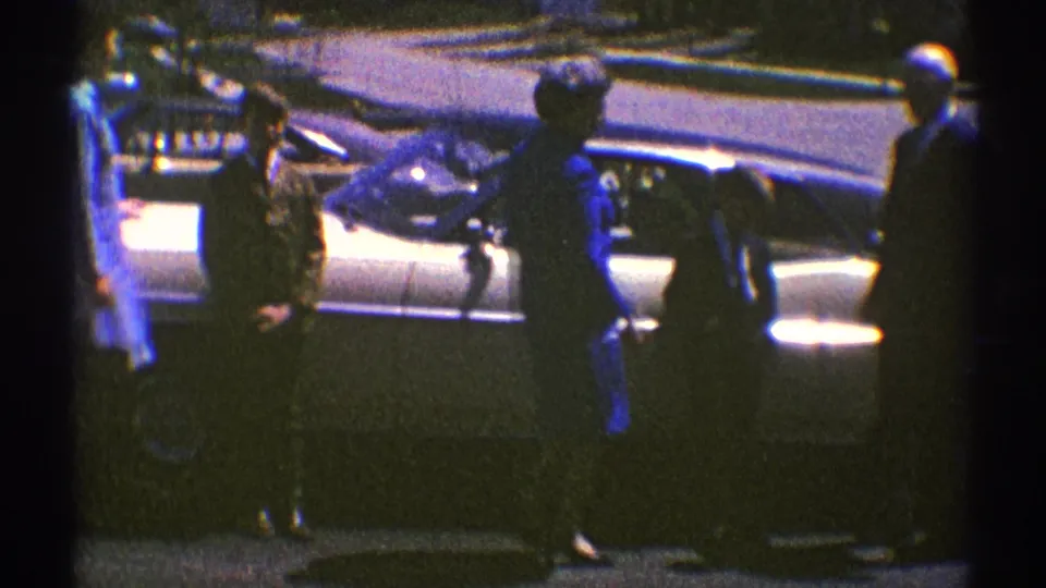 The 1969 Illinois home-movie frame shows adults loosely posing near a sedan, including a woman with a handbag in the foreground.