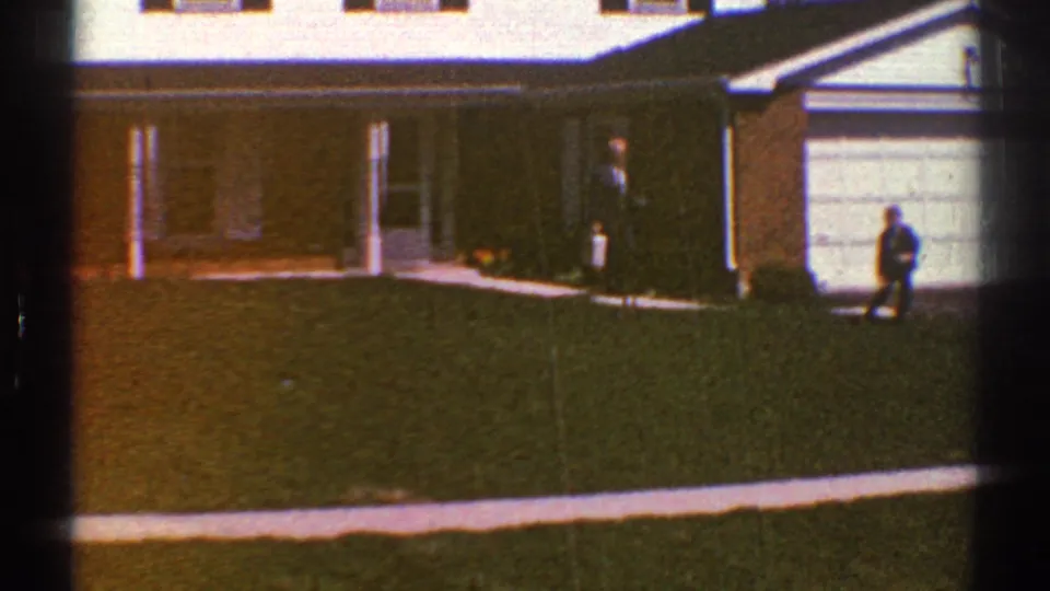1969 Clarendon Hills home-movie frame showing a suburban house, green lawn, concrete walk, garage door, and two distant people.