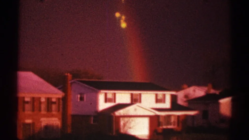 1969 home-movie frame of suburban Clarendon Hills houses, soft-focused and tinted by twilight, with a faint rainbow above.
