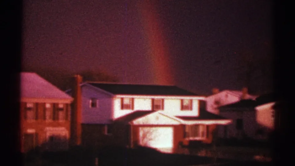 1969 8mm frame of a rainbow over a suburban block, with white siding, brickwork, roofs, and lawns visible.
