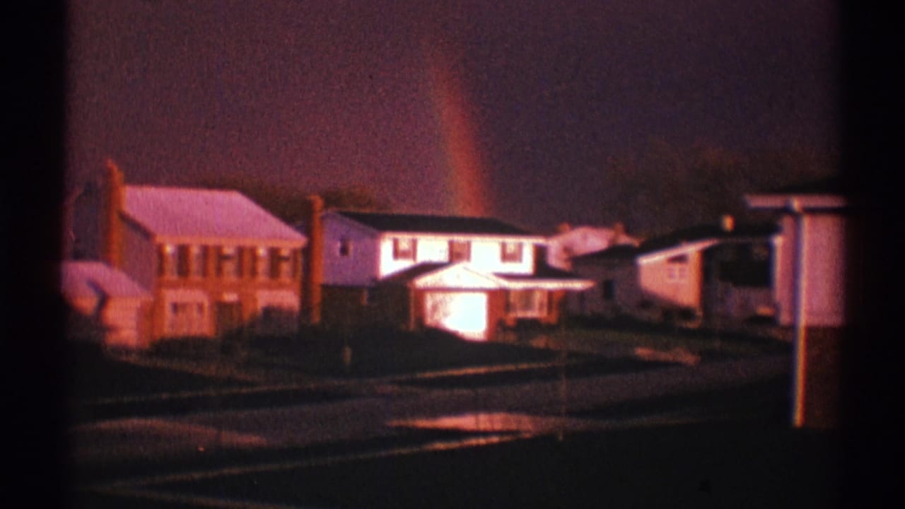 1969 Clarendon Hills home-movie frame with a rainbow, detached houses, glowing windows, wet street reflections, and dark clouds.