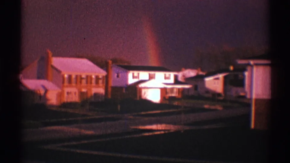 1969 Clarendon Hills 8mm frame showing a wet suburban street, detached houses, a roadside post, and a rainbow in a dark sky.