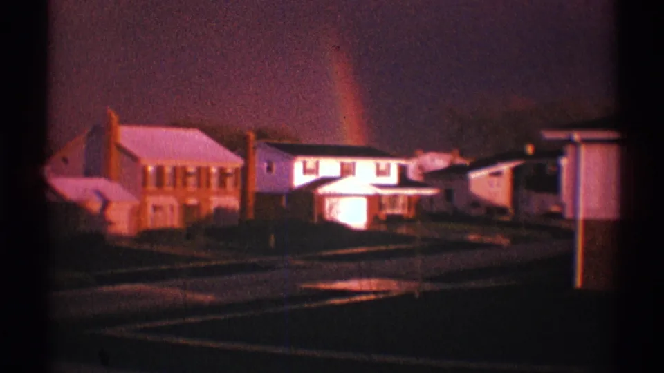 1969 Clarendon Hills home-movie frame with detached houses, a wet street, dark sky, and a rainbow partly visible behind rooftops.