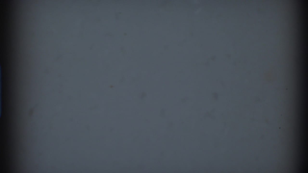 Low-light 8mm frame showing mostly soft gray-blue density, a dark vertical film edge, and scattered dust or emulsion marks.