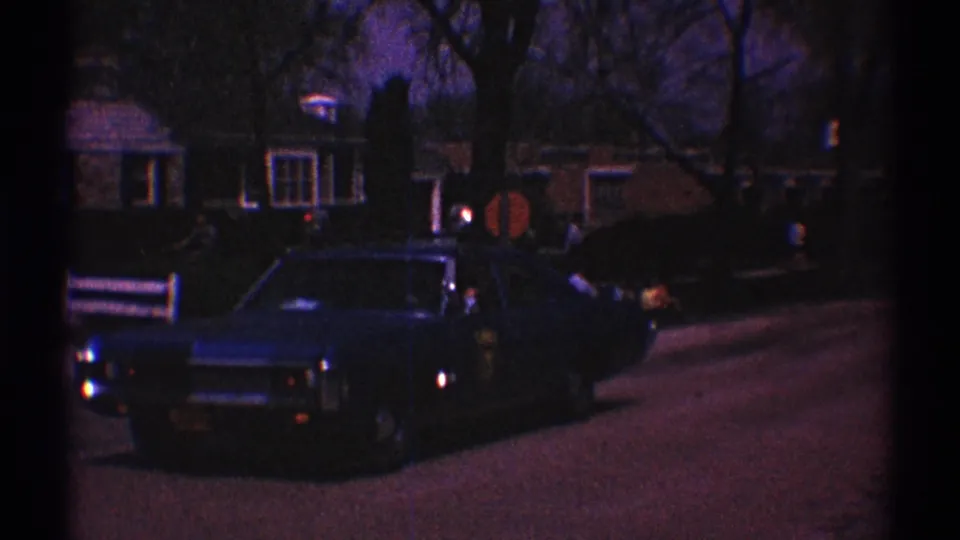 1969 Clarendon Hills home-movie frame showing a dark police car on a residential road at dusk, with houses, bare trees, and indistinct people nearby.