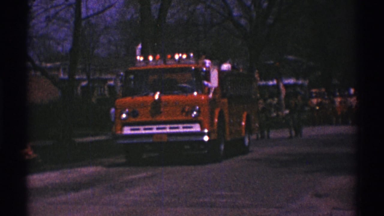 Orange fire truck with roof lights approaches on a dim residential street.