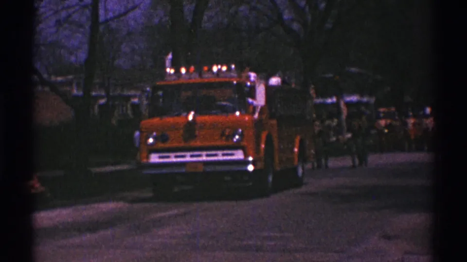 In Clarendon Hills, Illinois, an orange fire truck with lights on approaches along a suburban street in 1969 home-movie footage.