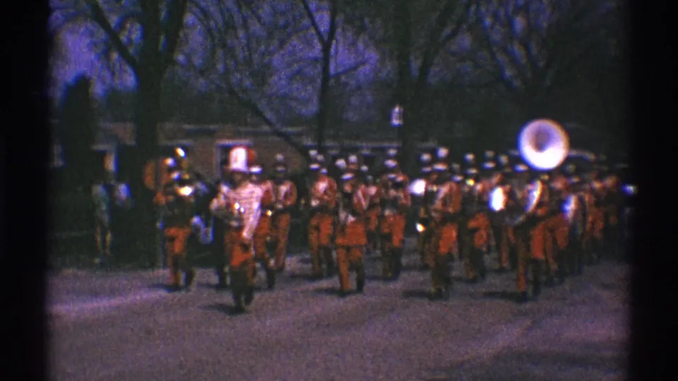 1969 Clarendon Hills home-movie frame showing a red-uniformed marching band advancing toward the camera on a tree-lined street.