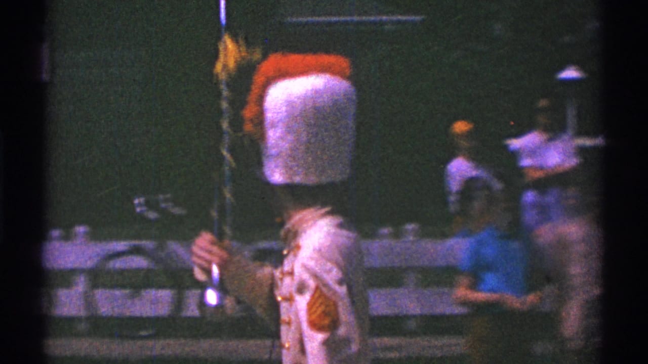 In 1969 Clarendon Hills parade footage, a young drum major in a tall plumed hat and red-and-white uniform holds a baton near spectators.