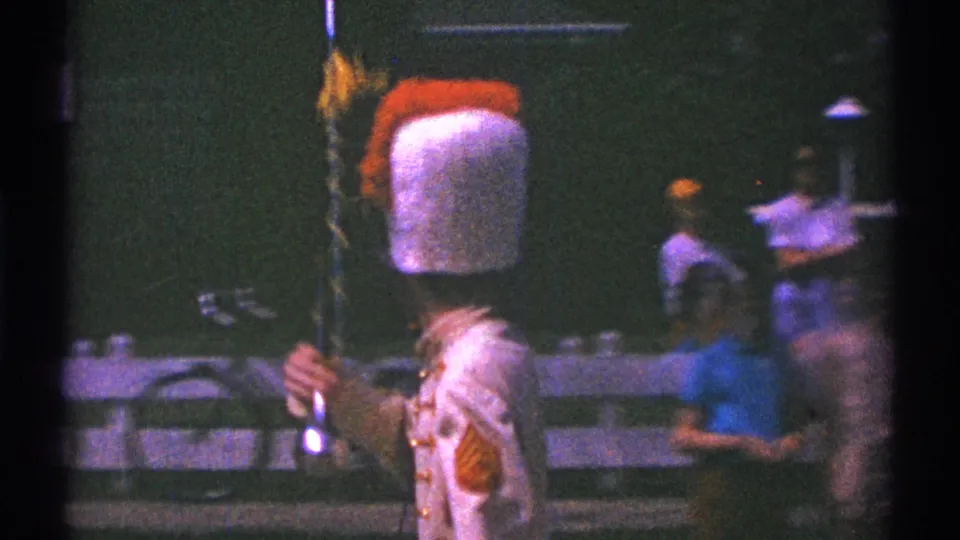 In 1969 Clarendon Hills parade footage, a young drum major in a tall plumed hat and red-and-white uniform holds a baton near spectators.