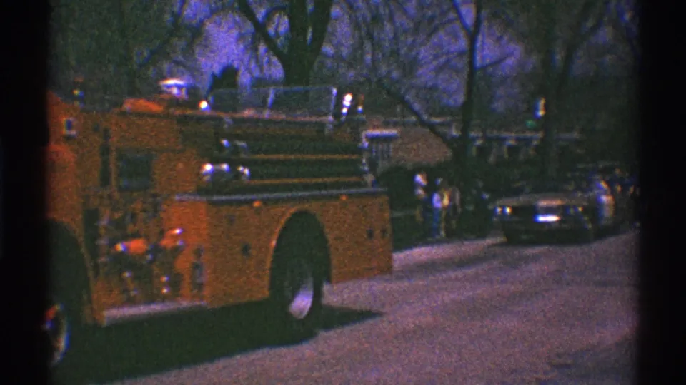 The rear side of the red fire truck moves past parked cars and distant onlookers on a Clarendon Hills residential street.