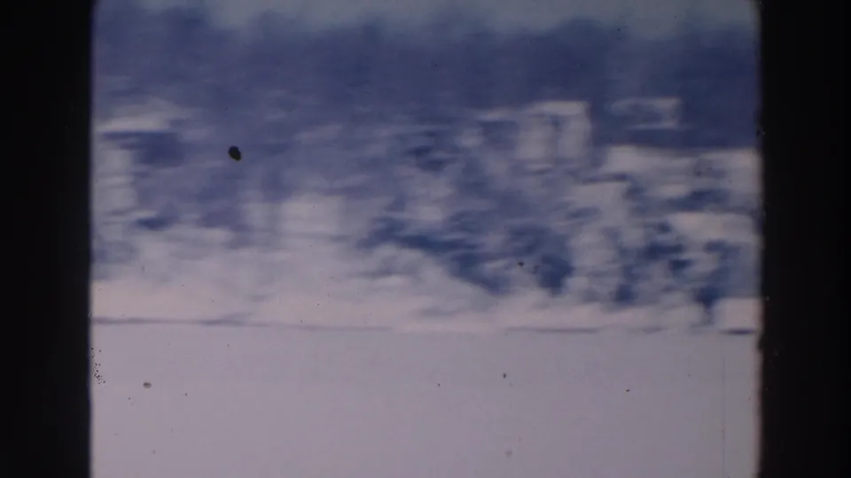 Soft 1964 8mm frame from Tustin, Wisconsin, showing snow, distant buildings, and a dark wooded horizon.