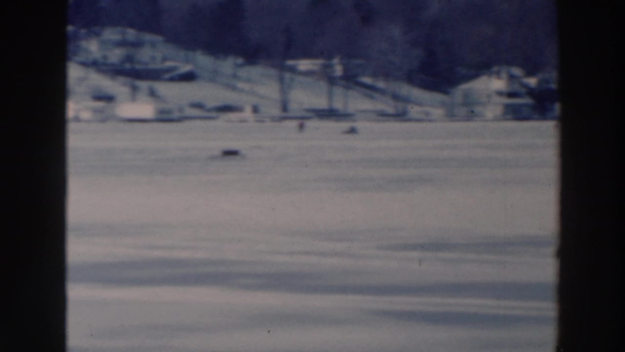 Two distant people walking on a snowy frozen lake with houses and trees along the shore.