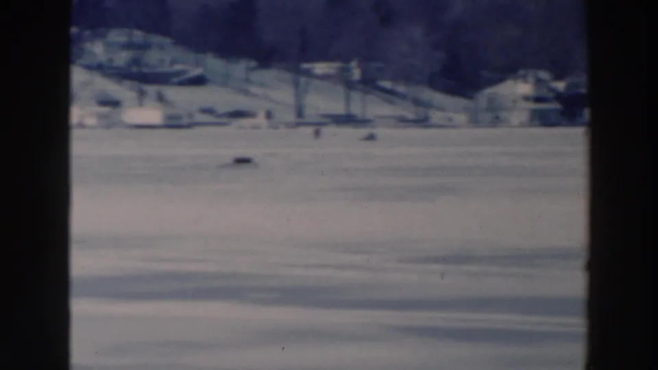 Two bundled people walk across a snowy frozen lake in Tustin, Wisconsin, with a distant shoreline of houses and winter trees.