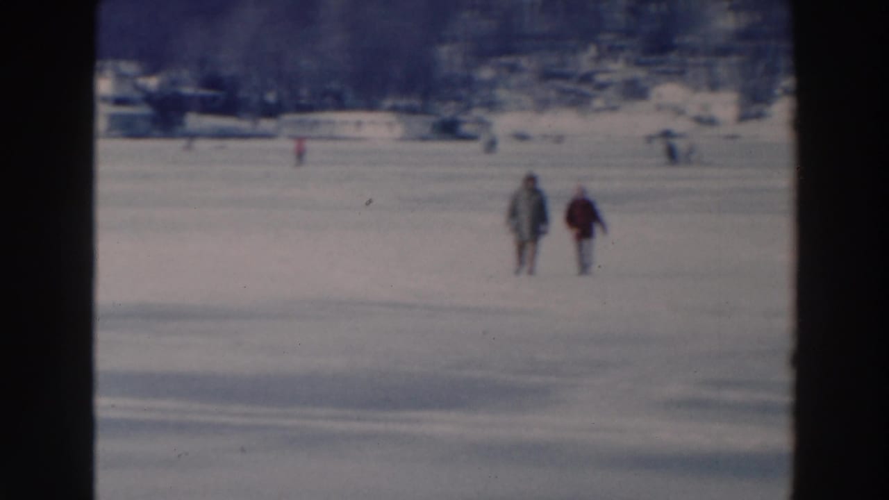 Two bundled figures move across the snowy ice with scattered people and buildings in the distance.