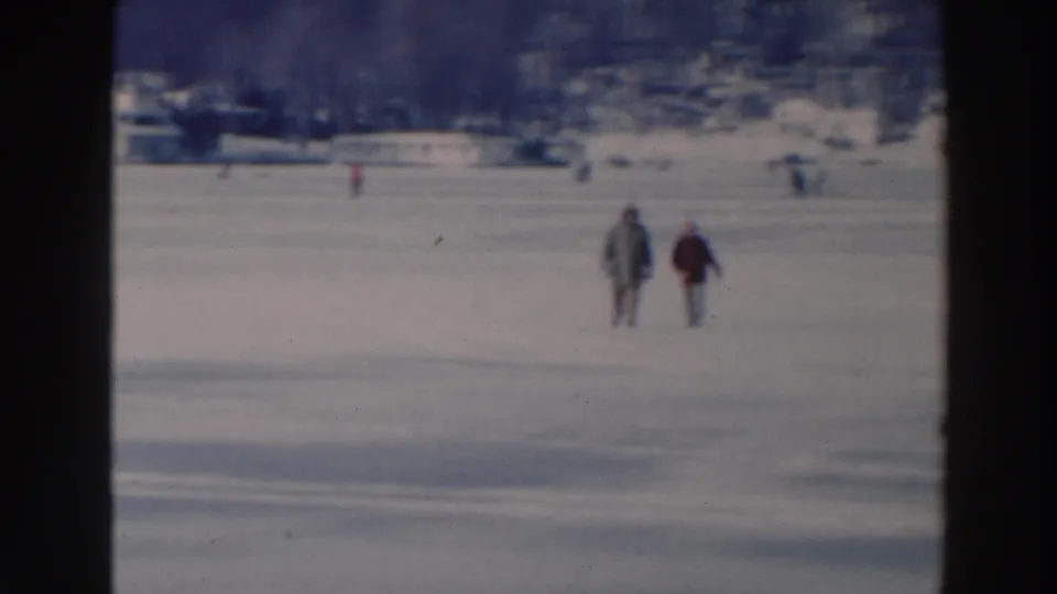 Two people in winter coats cross the frozen lake in the 1964 Tustin, Wisconsin home movie, with distant figures and shoreline buildings behind them.