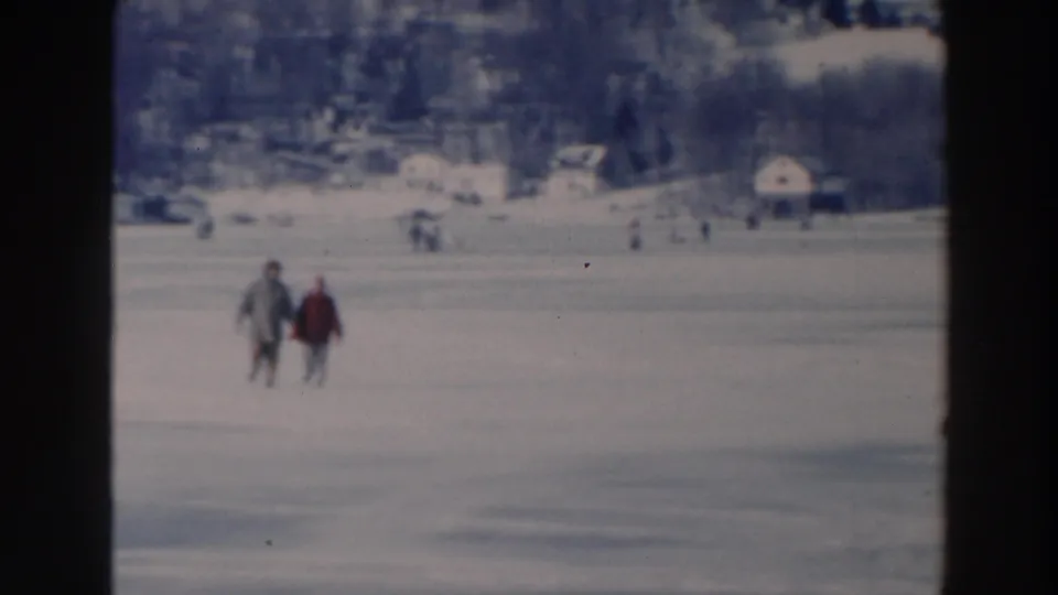 A pair of bundled walkers appears closer in the lower frame, crossing the frozen lake near Tustin, Wisconsin, with houses and trees beyond.