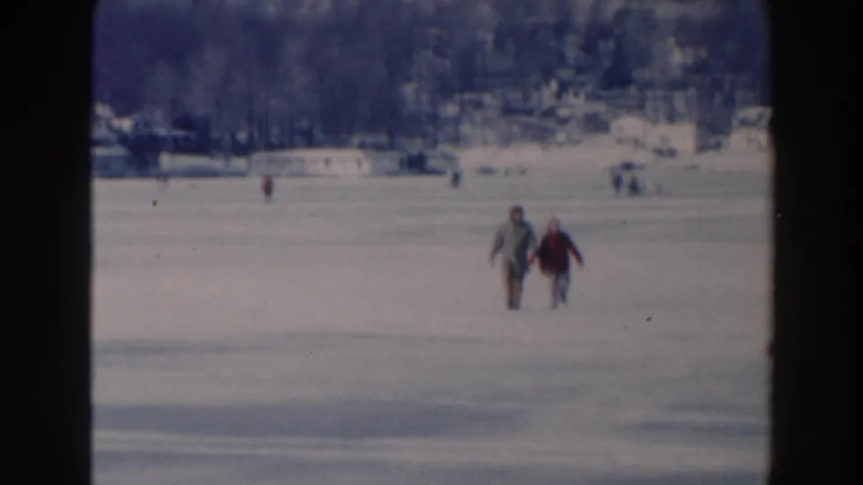 Two bundled people cross the snow-covered ice in Tustin, Wisconsin, framed by a broad frozen lake and distant winter shoreline.