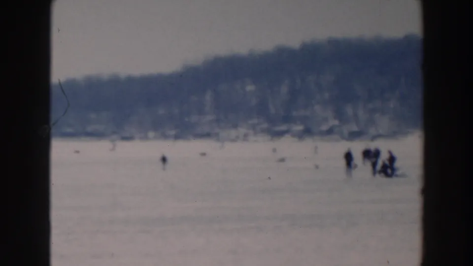 1964 Tustin, Wisconsin frame showing a distant group on the frozen lake below a snowy wooded shoreline.