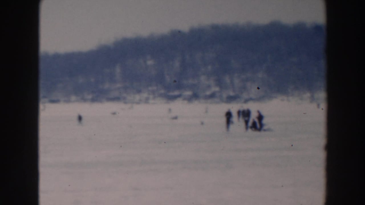 Distant people on a frozen lake with snowy ice and a dark treeline beyond.