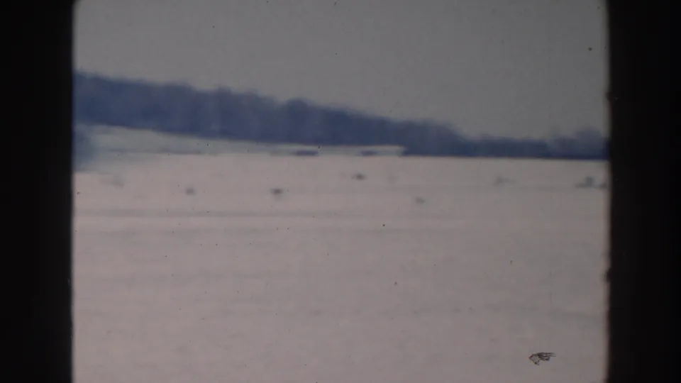 Wide home-movie view of a frozen Wisconsin lake with a low treeline and scattered distant activity on the ice.