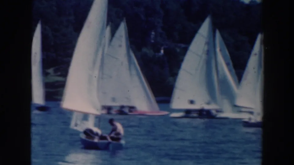 1964 Tustin, Wisconsin frame with a young man in a small boat near a cluster of white-sailed dinghies on the lake.