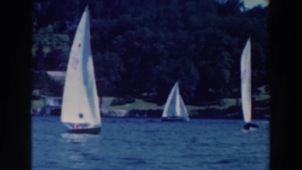 1964 Tustin, Wisconsin 8mm frame showing small sailboats spaced across the lake with trees and lakeside structures behind them.