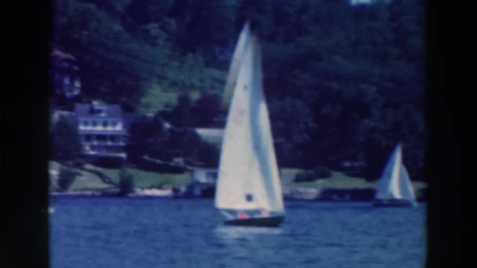 A white sailboat crosses blue water near a wooded shoreline in 1964 Tustin, Wisconsin home-movie footage.