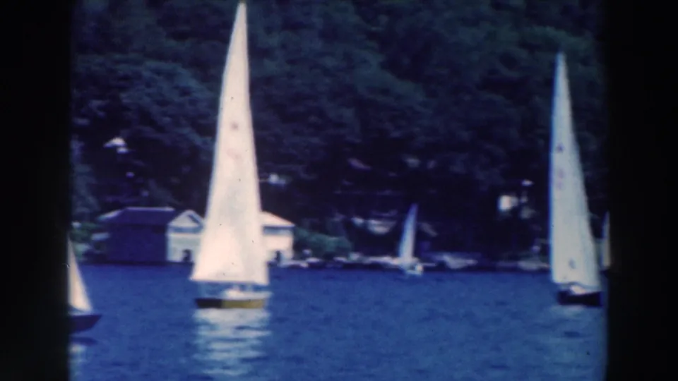 White sailboats sail past a wooded lakefront with a small boathouse or lakeside structure in 1964 footage.