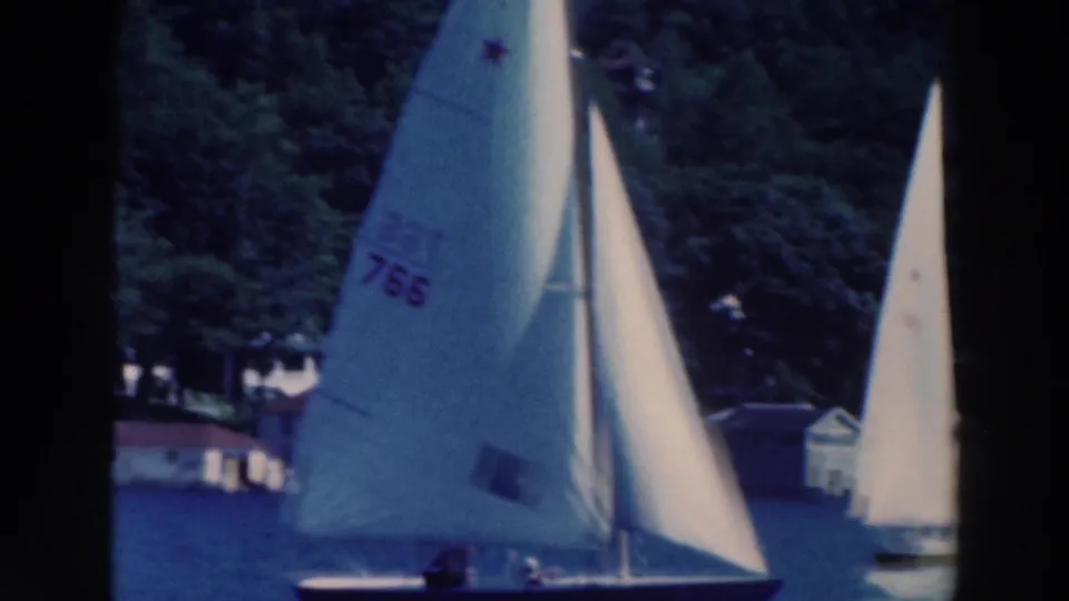 A numbered white sailboat passes close to the camera on a lake near other sailboats and a wooded Wisconsin shoreline.