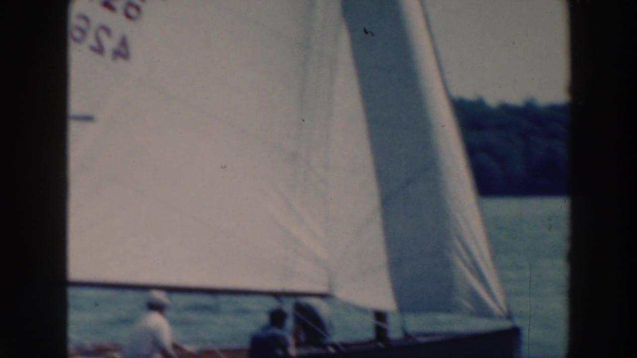 Close sailboat with a marked white sail and two people near the lower edge on a lake.