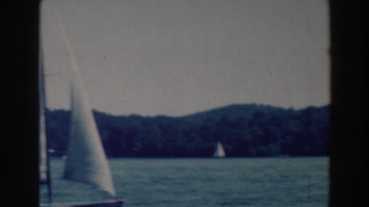 1964 Wisconsin lake sailing frame with one sailboat near center and another small sail farther along the forested shore.