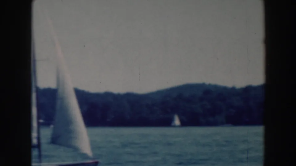1964 Wisconsin lake sailing frame with one sailboat near center and another small sail farther along the forested shore.