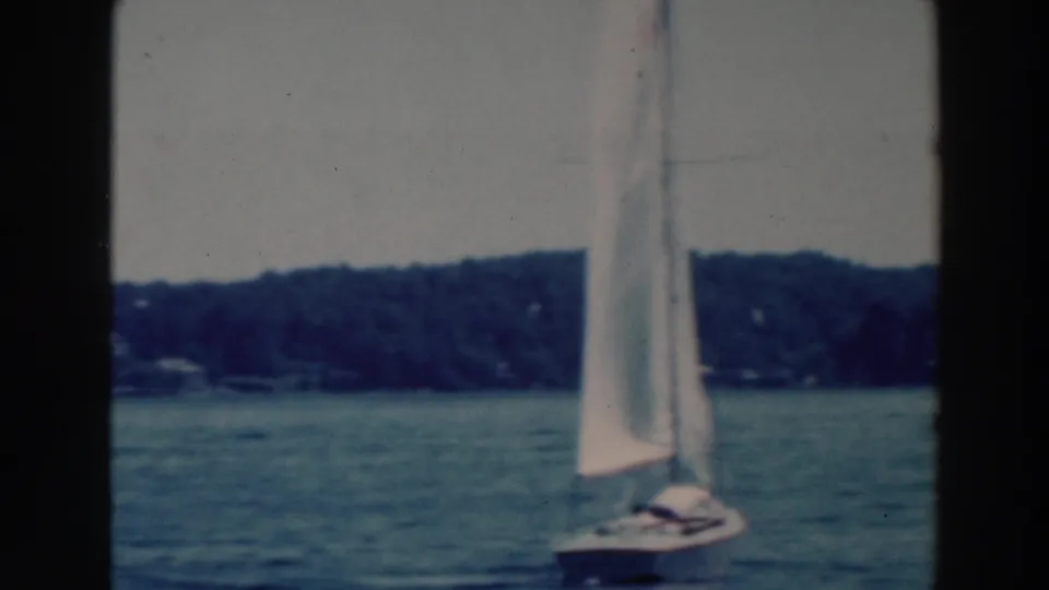 1964 Wisconsin lake scene with a small sailboat under white sails, distant wooded shore, and soft 8mm color.
