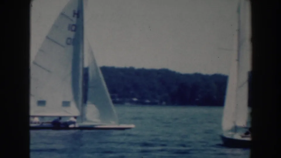 1964 Wisconsin home-movie frame of a marked sailboat on the lake, with another sailboat partly visible at right.