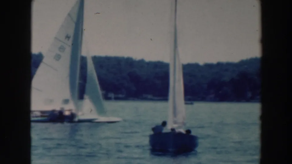 Home-movie frame from the 1964 Tustin, Wisconsin sailing clip showing small boats racing across a lake near a wooded shore.