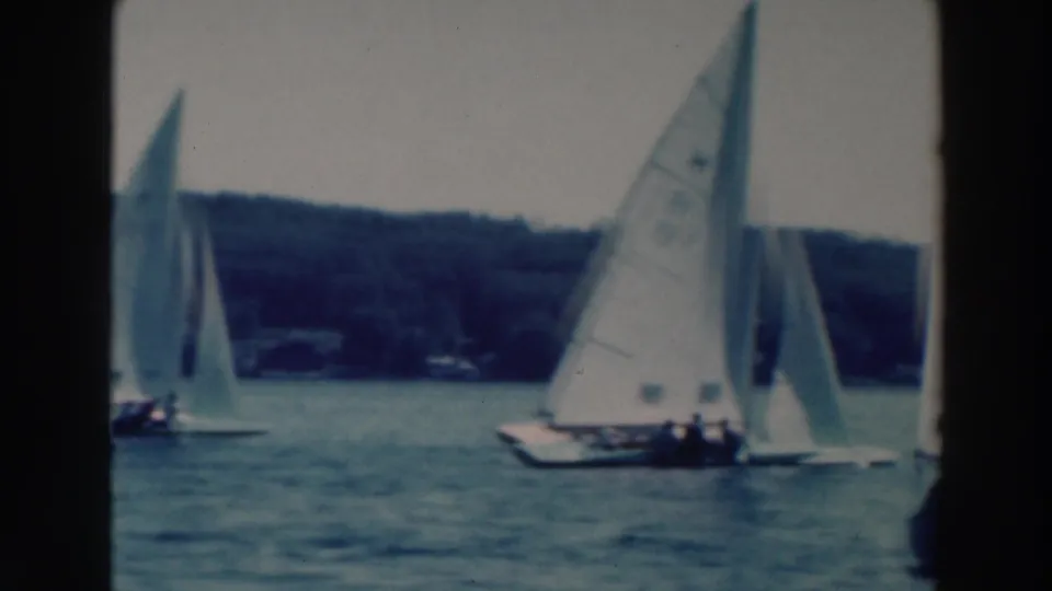1964 Tustin, Wisconsin lake race frame with a crewed sailboat in the foreground and other sails near the wooded shoreline.