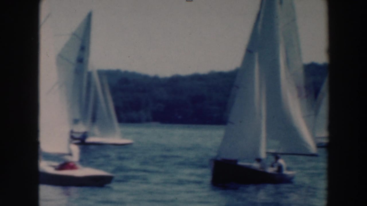 Sailboats pass at different distances on a lake, including one dark-hulled boat at right.