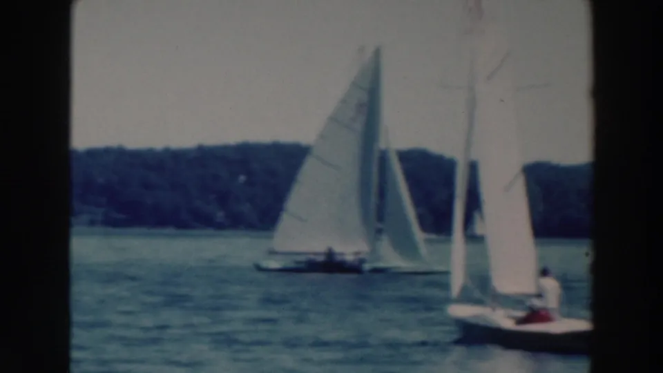 Late-frame view of the 1964 Tustin, Wisconsin sailing scene, with boats gliding past on rippled water and distant trees behind them.